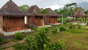A row of wooden cabins with thatched roofs, surrounded by lush greenery and a mountainous backdrop. - Balkondes Borobudur Duta Menoreh (Magelang)