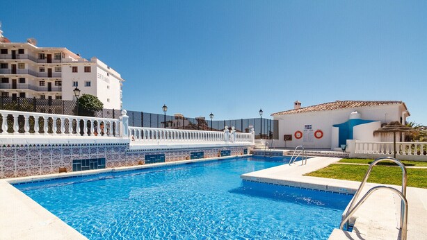En swimmingpool med ett vitt kaklat poolområde, en metallstege och en byggnad i bakgrunden med texten ”EJF FLUIDO”.
