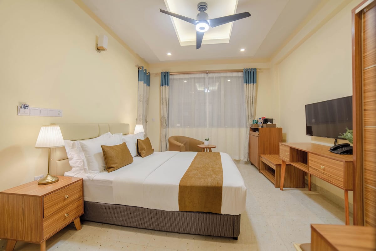 Habitación de hotel con cama, mesita de noche, lámpara, escritorio, silla, televisor y ventilador de techo.