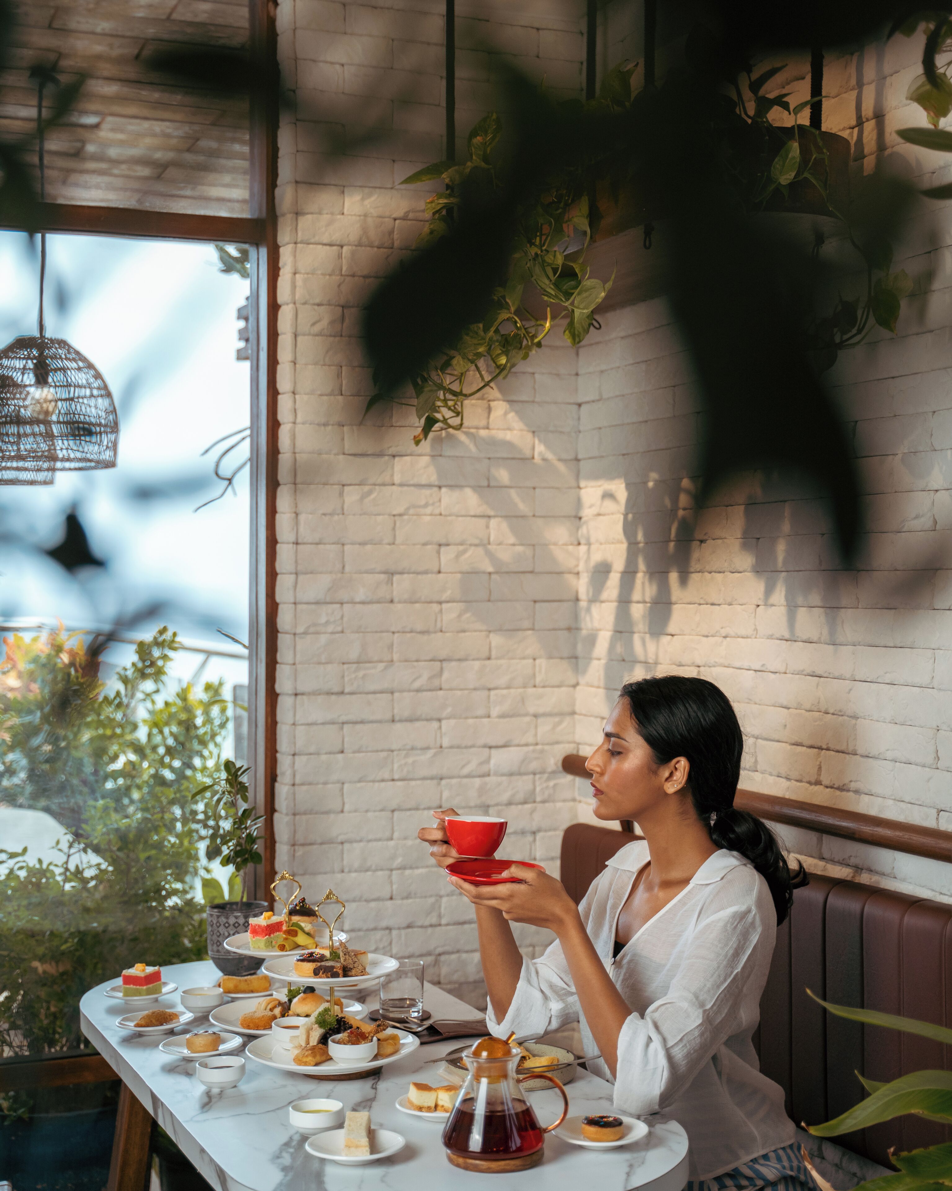 A woman seated at a table with various pastries and a teapot, in a room with a brick wall and hanging plants.