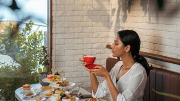 A woman seated at a table with various pastries and a teapot, in a room with a brick wall and hanging plants.