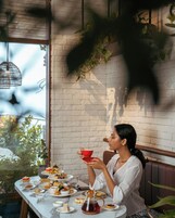 Una mujer sentada a una mesa con varios pasteles y una tetera, en una habitación con pared de ladrillo y plantas colgantes.