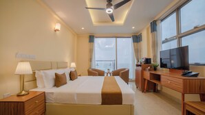 A hotel room with a bed, bedside tables, a desk with a TV, a ceiling fan, and large windows. - Amber Beach (Hulhumalé)