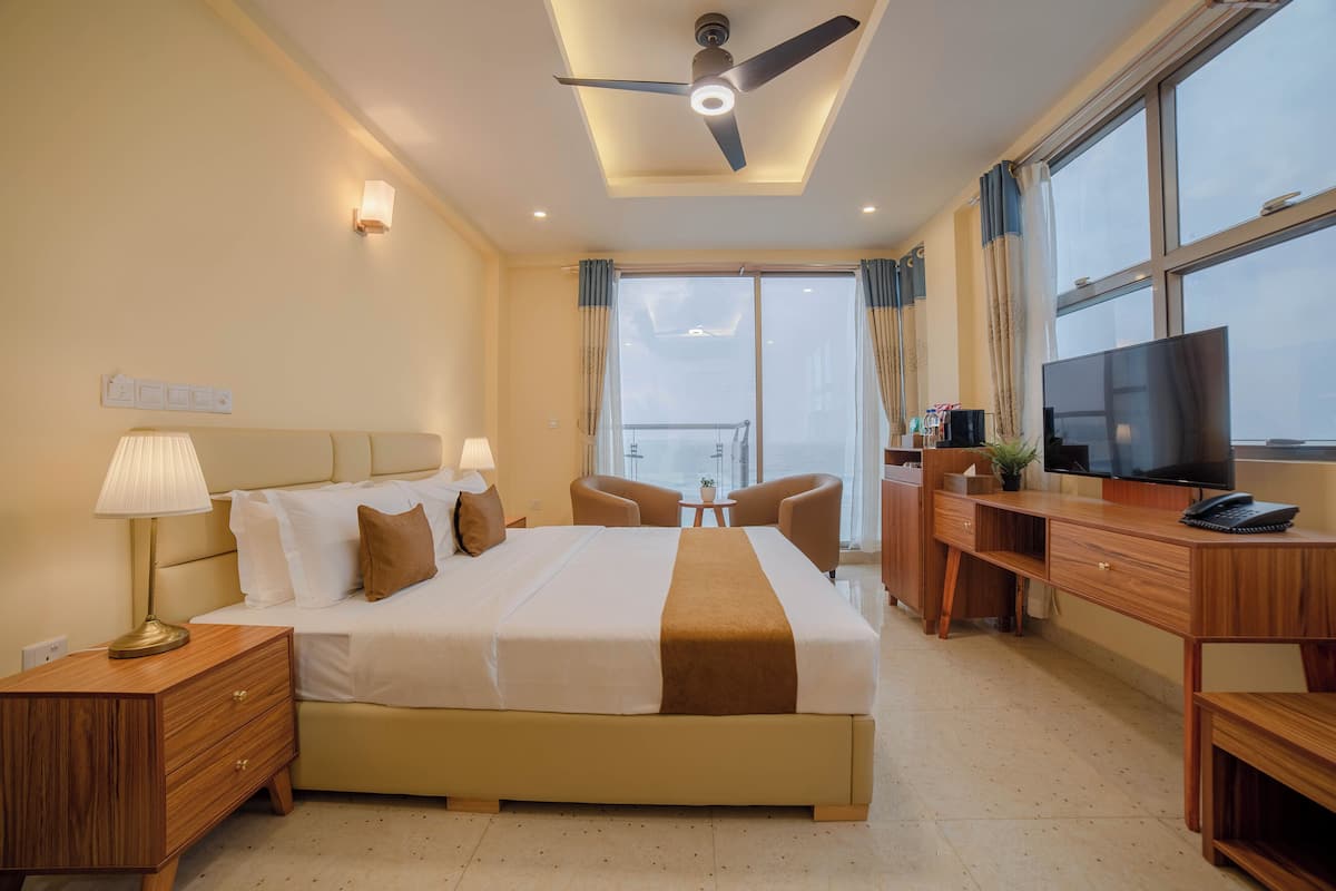 Habitación de hotel con cama, mesitas de noche, escritorio con televisor, ventilador de techo y ventanas amplias.