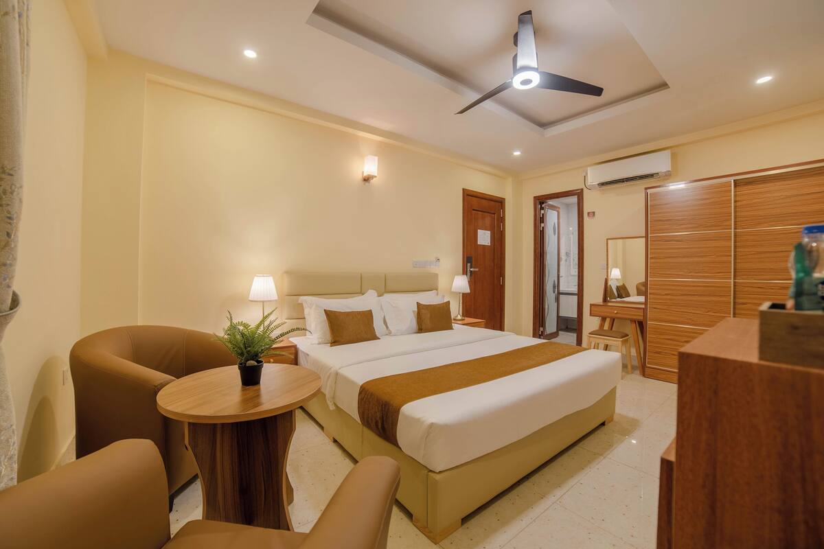 Habitación de hotel con cama, dos sillas, una mesita de noche con una planta, un escritorio con lámpara y un armario.