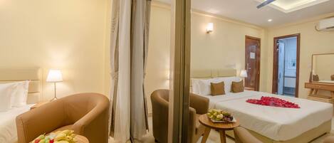 A hotel room with a bed, two armchairs, a small table with fruit, a ceiling fan, and a nightstand with a lamp.
