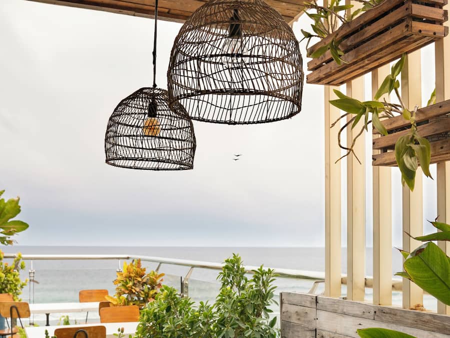Un área de comedor al aire libre con vistas al mar, que cuenta con una mesa de madera, sillas, lámparas colgantes de mimbre y plantas en macetas.