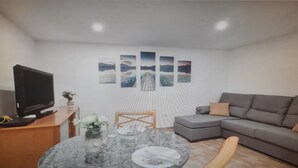 A living room with a grey sofa, a TV on a wooden stand, a glass coffee table with a vase and papers, and a wall with framed pictures. - Apartment in Tenerife South (Cabo Blanco - Arona)