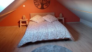 A bedroom with a sloped ceiling, a bed with patterned bedding, two bedside tables with lamps, and a wall mural of a tree. - Comfortable cottage "Les Délices" (Guewenheim)