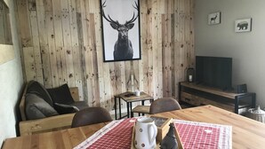 A cozy room with a wooden wall, a large framed deer picture, a TV, and a dining table with a checkered placemat. - Comfortable cottage "Les Délices" (Guewenheim)