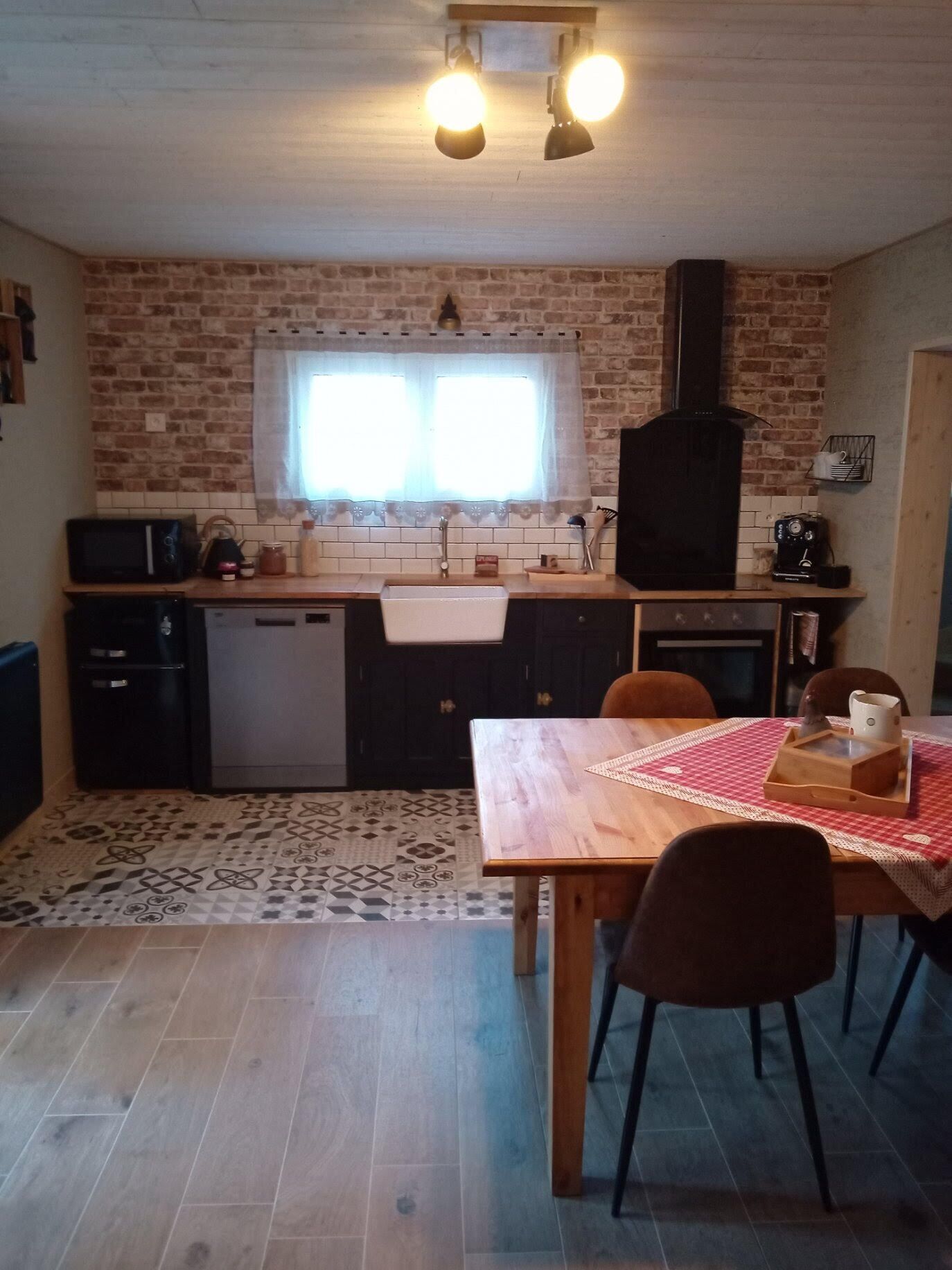 Une cuisine de taille modeste avec un mur en briques, une fenêtre avec des rideaux, un évier, une cuisinière et un coin repas comprenant une table et des chaises.