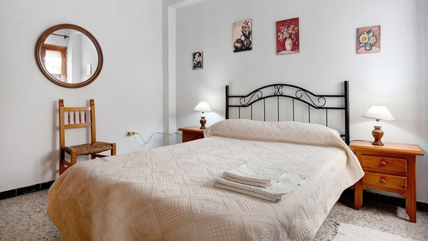 A bedroom with a bed, a wooden chair, a nightstand with a lamp, a mirror on the wall, and three framed pictures.