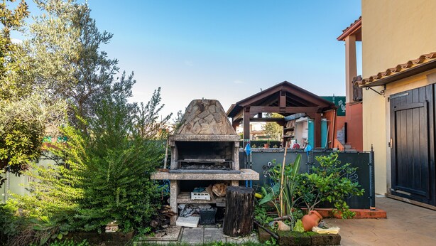 Ein Steinofen im Freien, eine Holzpergola und ein Haus mit Ziegeldach.