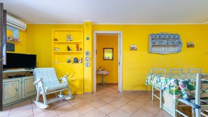 Ein Zimmer mit gelben Wänden, einem Fernseher, einem Schaukelstuhl, einem Esstisch mit Stühlen und Regalen mit Dekoration.