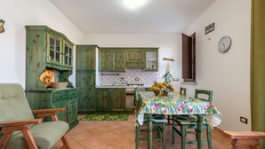 Une cuisine avec des meubles de rangement verts, une table à manger recouverte d’une nappe à motifs floraux et une chaise.