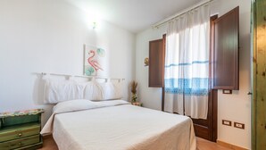 Une chambre à coucher avec un lit, une table de chevet, une commode verte, un tableau, une fenêtre avec des rideaux et une porte.