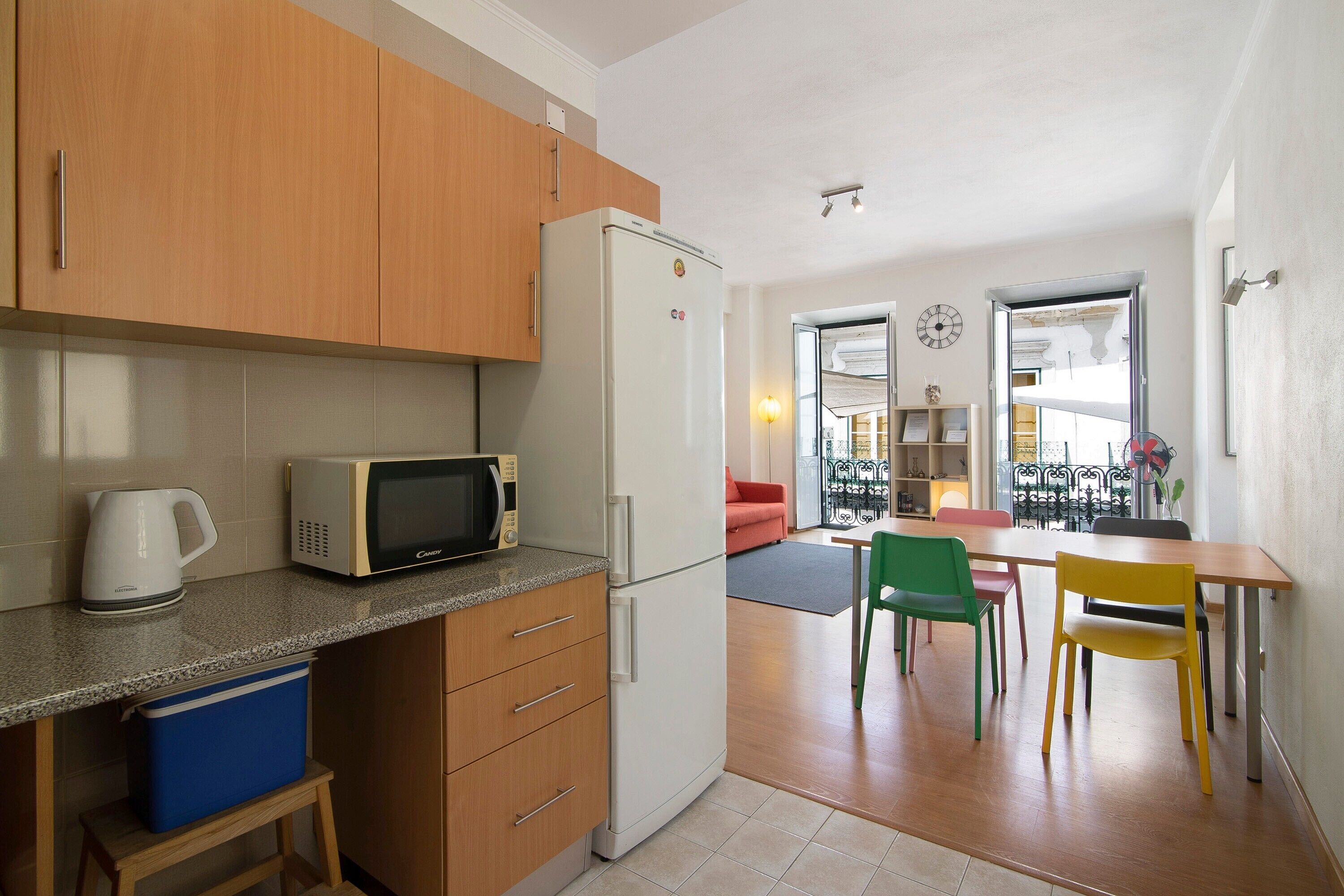 A compact kitchen with a refrigerator, microwave, and kettle, a dining area with chairs, and a visible clock on the wall.