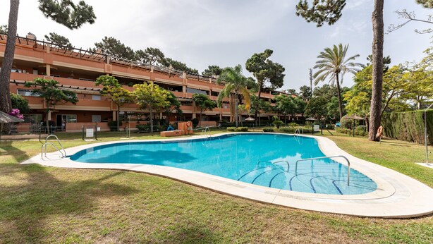 Una piscina rodeada de césped, árboles y un edificio de varios pisos con balcones.