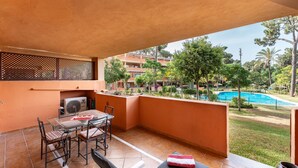 Un balcón con mesa y sillas, un sofá y vistas a una piscina y zonas verdes.