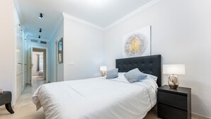 Un dormitorio con cama, mesitas de noche, un banco y un pasillo que conduce a otra habitación.