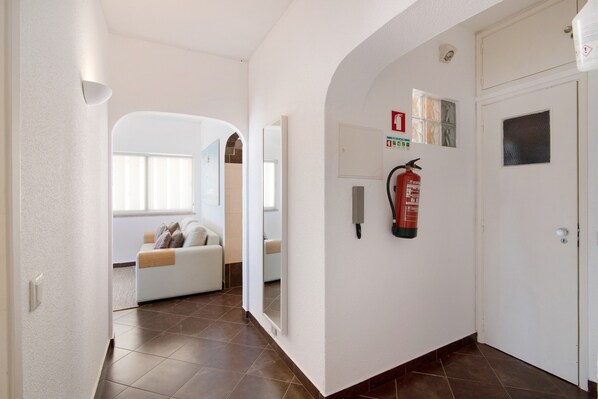 A hallway with a fire extinguisher mounted on the wall, a white door, and a seating area with a sofa and cushions.
