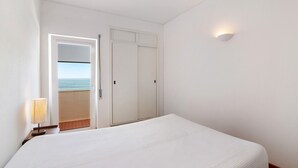 A hotel room with a bed, a nightstand, a lamp, a door leading to a balcony with a view of the sea, and a wall-mounted light fixture. - Ocean View Apartment with Sea View, Wi-Fi (Armação de Pêra)