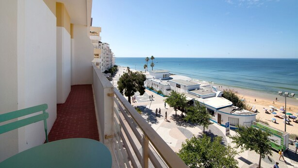 A balcony with a view of a beach, a parking area, and a building with a white facade. - Ocean View Apartment with Sea View, Wi-Fi (Armação de Pêra)