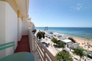 A balcony with a view of a beach, a parking area, and a building with a white facade.
