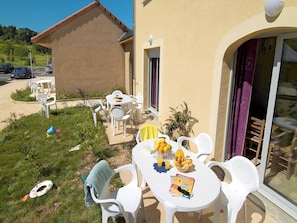 Eine Terrasse im Freien mit weißen Plastikstühlen und -tischen, ein kleines Gebäude im Hintergrund und ein geparktes Auto in der Nähe.