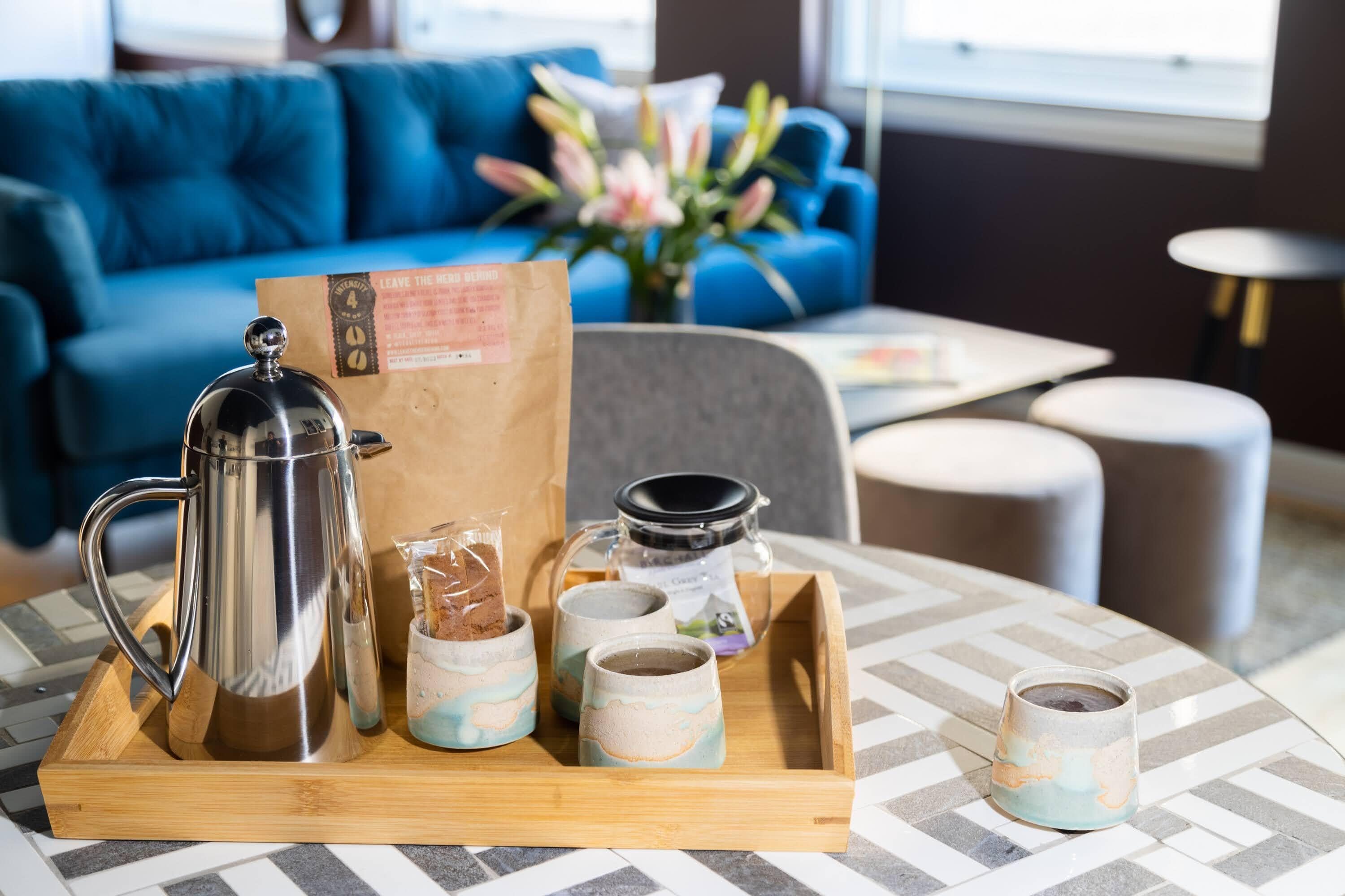 A tray with a coffee pot, two mugs, a jar, and a paper bag on a table in a modern room with a blue couch and a vase of flowers.