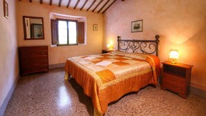 Una camera da letto con un letto, una testiera in legno, un comodino, una cassettiera, una finestra e uno specchio fissato alla parete.