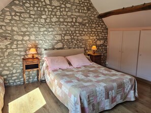 A bedroom with a stone wall, a bed with a quilt, two bedside lamps, a wooden nightstand, and a wardrobe.