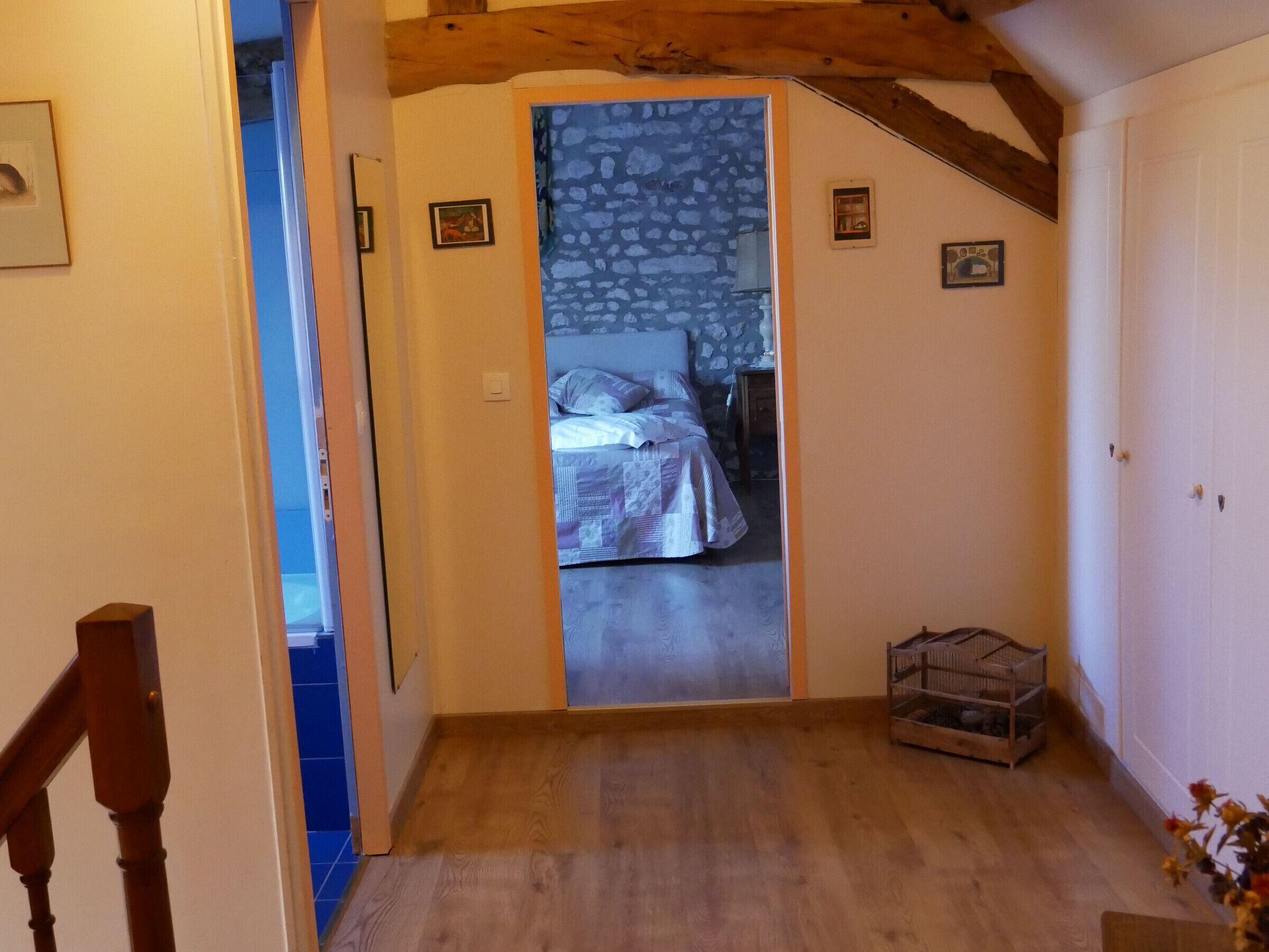 Un couloir étroit mène à une chambre dont un des murs est en pierre, qui contient un lit en bois et une petite caisse en bois.