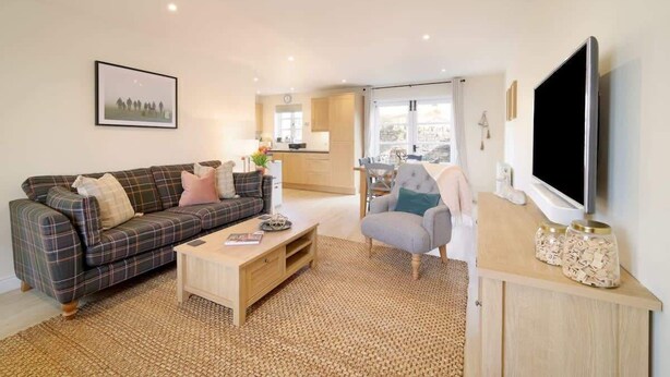 A living room with a plaid sofa, a wooden coffee table, a grey armchair, a flat-screen TV, and a kitchen in the background. - Townhouse with Views of Wells Cathedral (Wells)