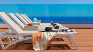 Une terrasse baignée de soleil, avec des chaises longues blanches, une table sur laquelle reposent un chapeau et des lunettes de soleil, et un verre de vin.