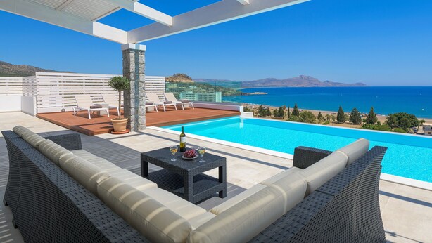 Un espace piscine extérieur moderne offrant une vue dégagée sur la mer, agrémenté d’un pilier en pierre et d’un coin salon avec une table et des boissons.