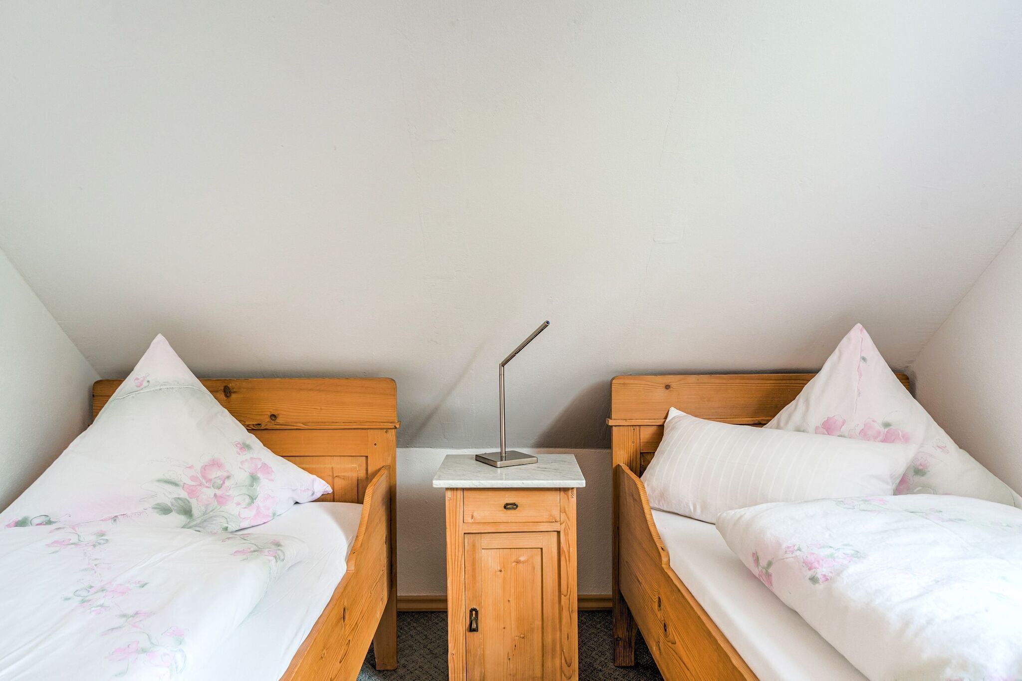 A small, cozy room with two wooden beds, each with floral bedding, a nightstand with a lamp, and a small wooden cabinet.