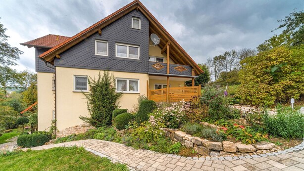 A two-story house with a balcony, surrounded by a well-maintained garden with various plants and a stone pathway. - Apartment in Marktrodach mit Sauna (Marktrodach)