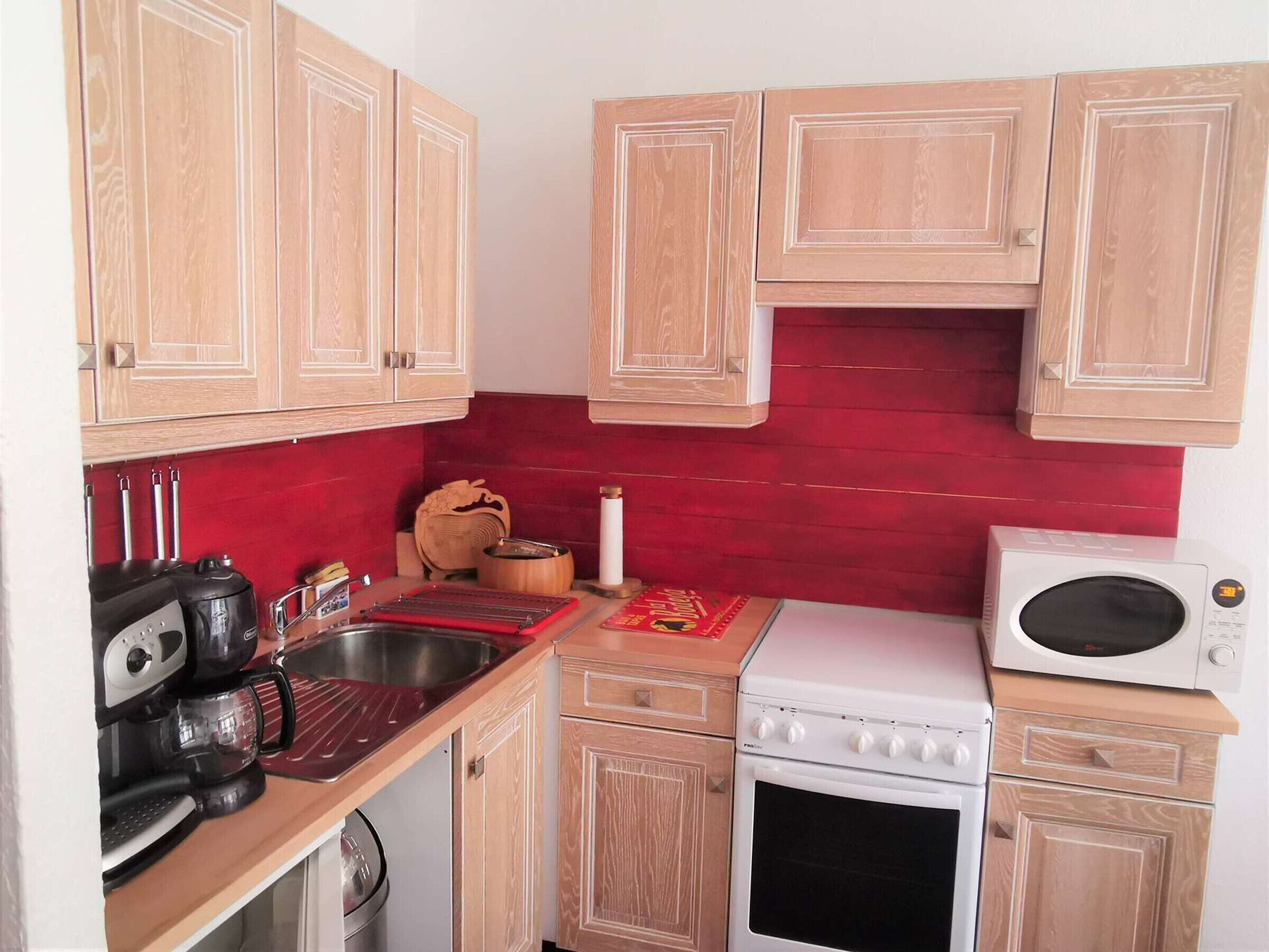 A compact kitchen with wooden cabinets, a red backsplash, a microwave, a stove, and a coffee maker.