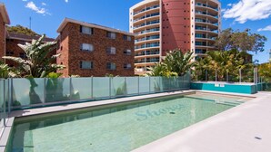 A swimming pool with a clear blue water surface, surrounded by a glass fence, and adjacent to a brick building with multiple windows.