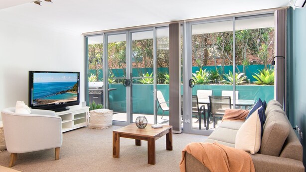 A modern living room with a television, comfortable seating, and a view of a garden through large sliding glass doors.