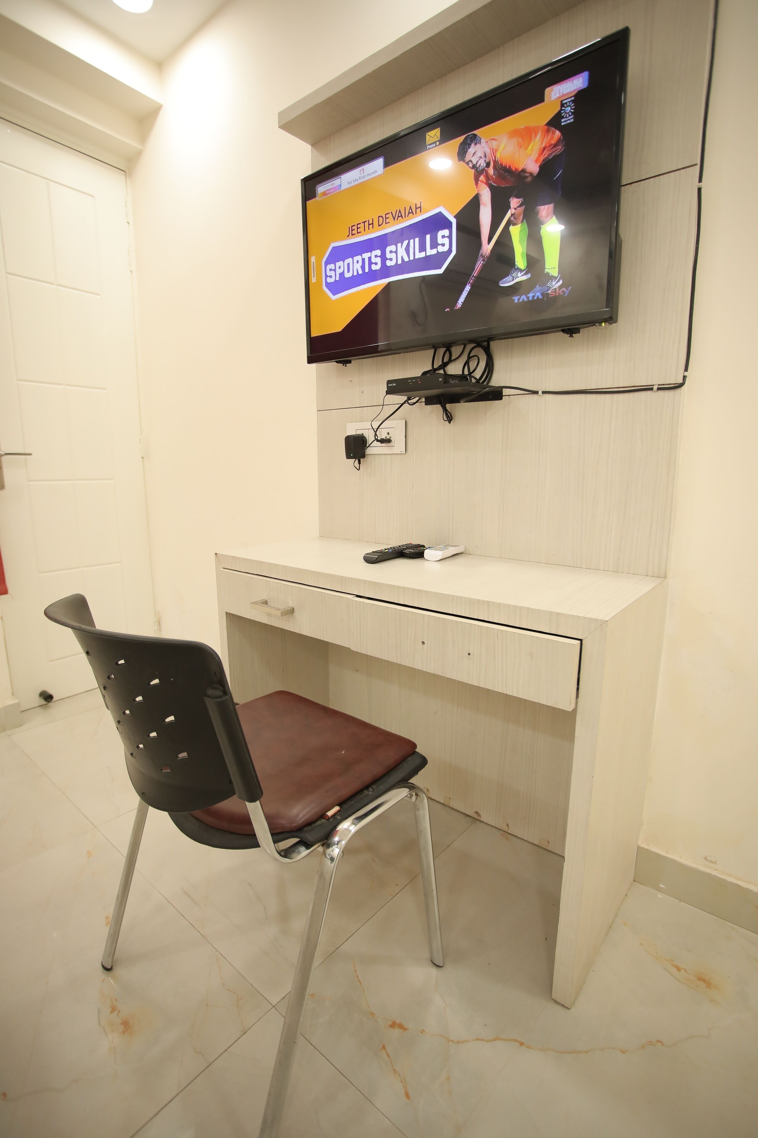 Habitación con una televisión instalada en la pared mostrando un anuncio de habilidades deportivas, un escritorio con una silla y una puerta blanca.