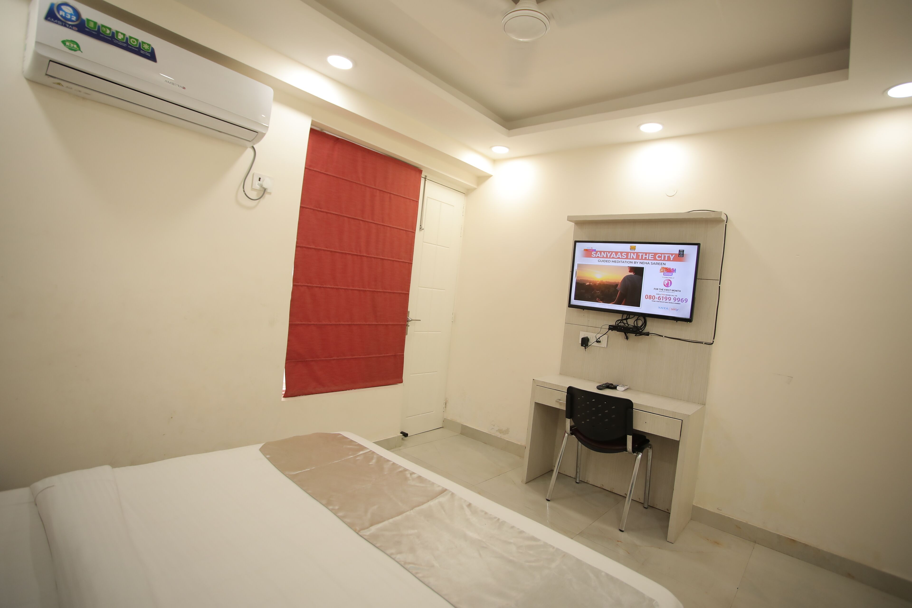 A hotel room with a bed, a desk, a chair, an air conditioning unit, a television mounted on the wall, and a red curtain.