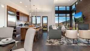 A modern kitchen with a central island, a dining area with a table and chairs, a living room with a sofa and armchairs, and large windows offering a view of the outdoors.