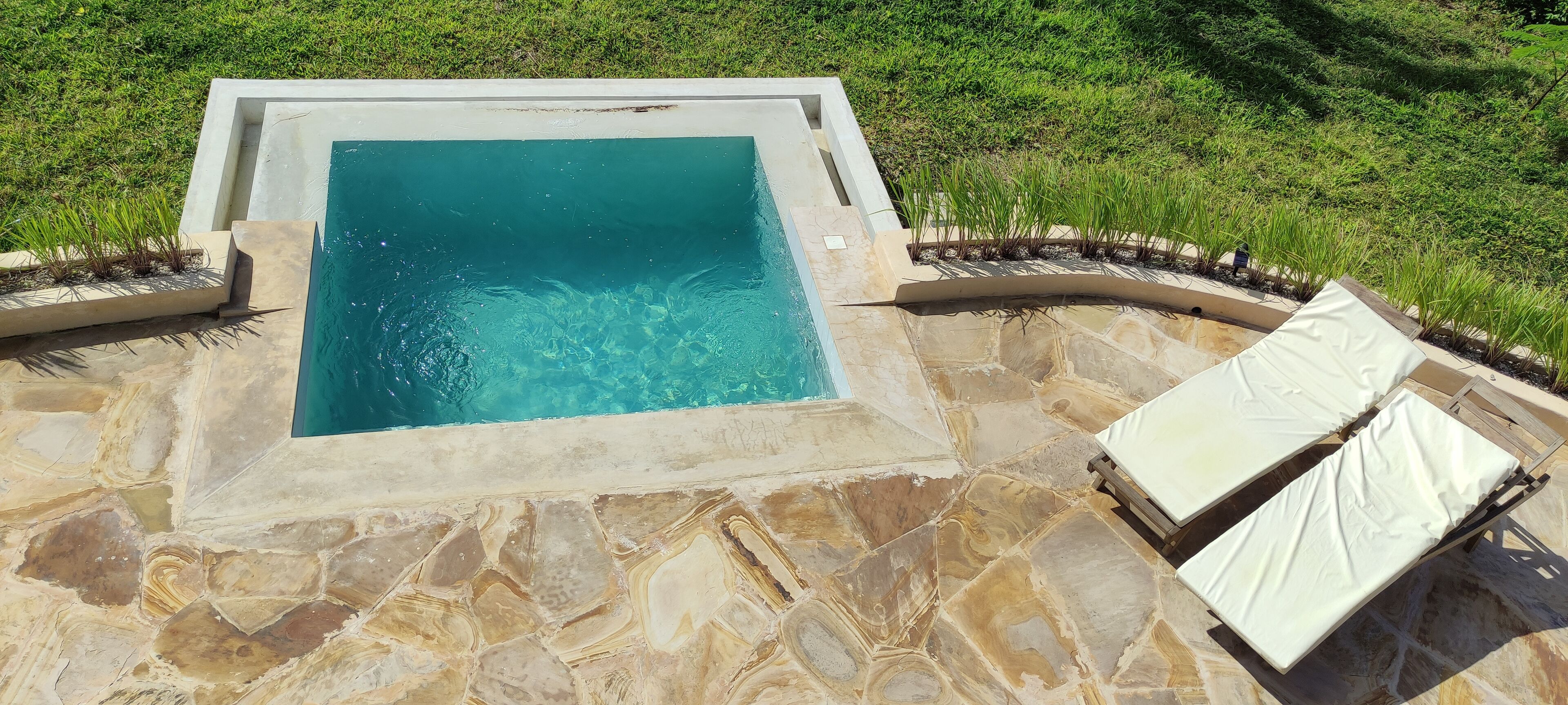 Una piscina rectangular con borde de piedra, rodeada por un patio de piedra. Hay dos sillones de descanso con cojines blancos.