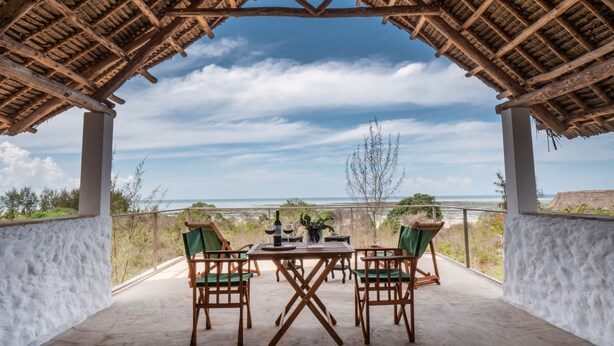 Um pátio ao ar livre com um telhado de palha, uma mesa com cadeiras e vista para o oceano.
