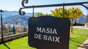 A sign reading "MASIA DE BAIX" against a scenic backdrop of mountains and autumn foliage.