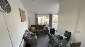 A modern living room with a grey sofa, a glass dining table, and a black clock on the wall. - Cosy 2 Bedroom Lodge with onsite Pool (Oulton Broad, Lowestoft)