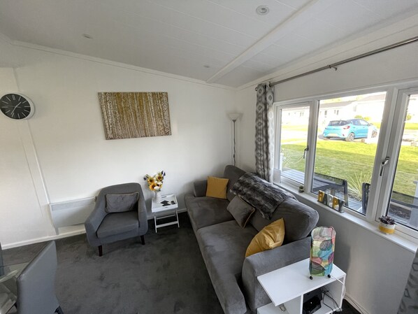 A modern living room with a grey sofa, armchairs, a small white table with a vase of flowers, and a window with a view of a blue car and greenery outside.