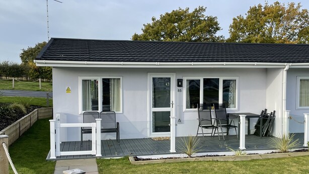 A small, single-story building with a dark roof and white exterior walls. It features a covered patio area with a table and chairs, and a small garden with plants and a wooden fence. - Cosy 2 Bedroom Lodge with onsite Pool (Oulton Broad, Lowestoft)